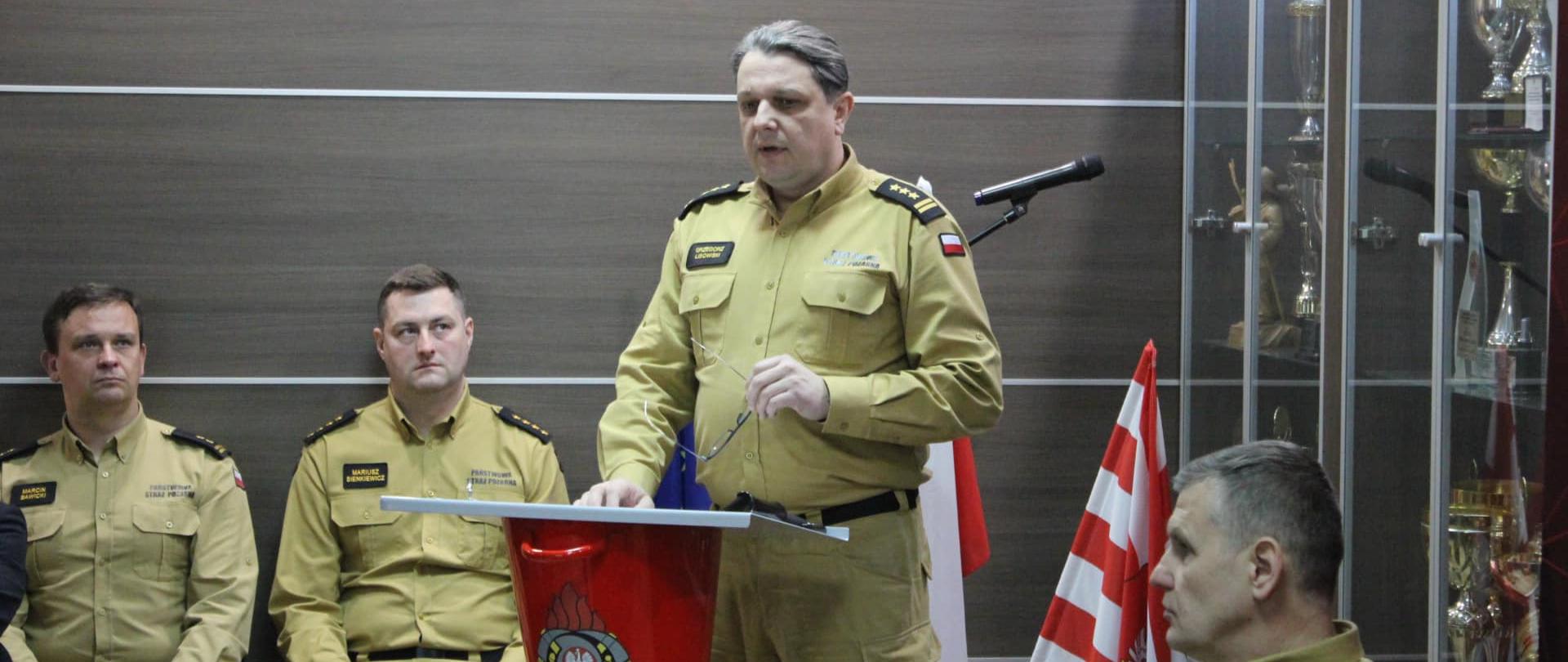 Narada roczna podsumowująca funkcjonowanie ochrony przeciwpożarowej w powiecie bielskim za rok 2024 - przemówienie st. bryg. Grzegorza Lisowskiego, w tle 3 funkcjonariuszy KP PSP w Bielsku Podlaskim