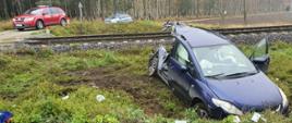 Rozbity samochód osobowy w kolorze niebieskim widziany od przodu wryty w glebę przy przejeździe kolejowym. W tle zdjęcia widać samochód strażacki i tory.
