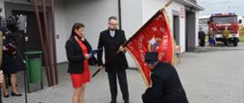 Strażak z OSP w Aleksandrowie klęka na jedno kolano i całuje nowy sztandar jednostki. Sztandar trzyma w rękach burmistrz miasta i gminy Wiskitki. Obok niego soi kobieta i trzyma w ręku dokument