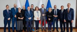 Zdjęcie zbiorowe, na tle białej ściany na której wisi pięć portretów w złoconych ramach na podłodze z drewnianych klepek stoi grupa 12 osób, pośrodku minister Kulasek, za nimi pod ścianą flagi Polski i UE.