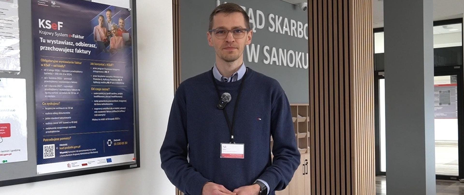 Pracownik Urzędu Skarbowego w Sanoku. Obok wisi plakat KSeF. Z tyłu na ścianie napis Urząd Skarbowy w Sanoku.