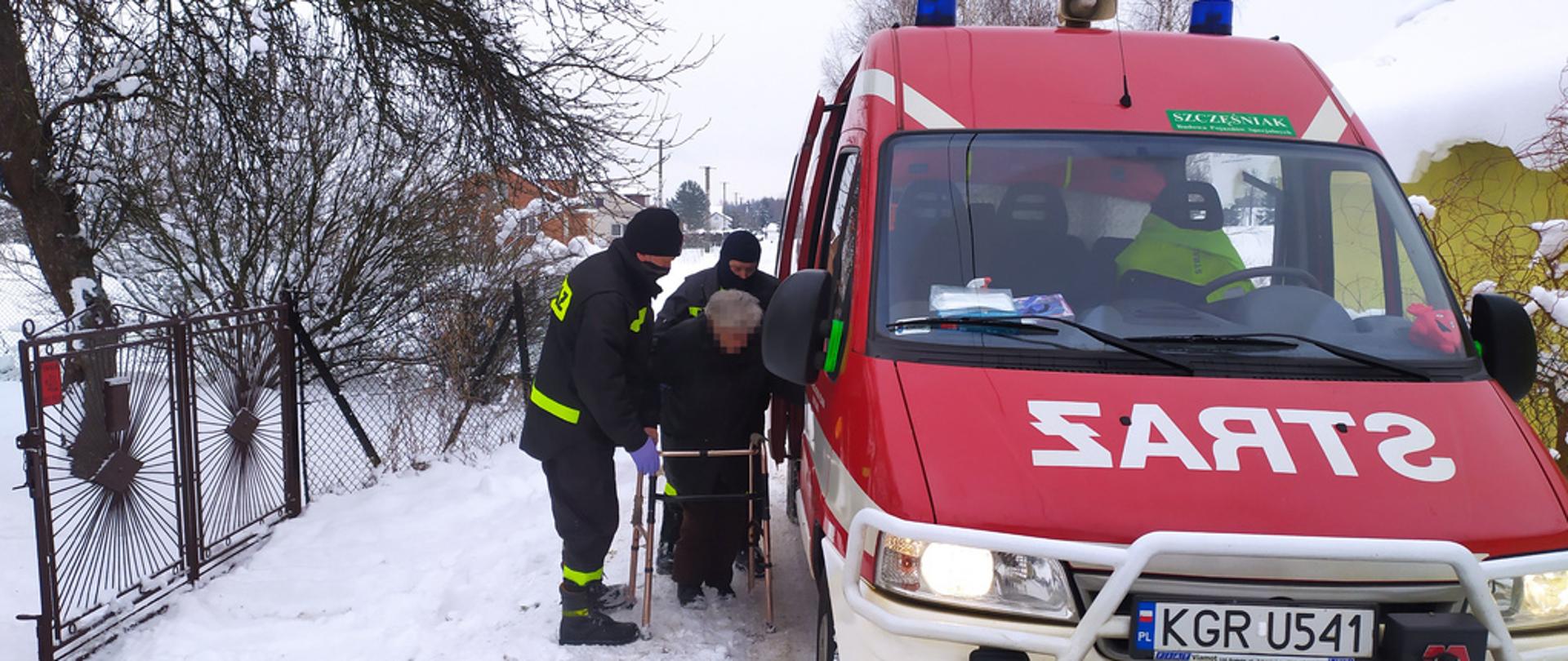 Dwóch strażaków na zdjęciu pomaga seniorce, która porusza się o balkoniku wsiąść do strażackiego samochodów w celu przetransportowania jej do punktu szczepień.