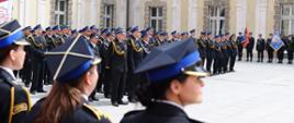 Strażacy PSP oraz funkcjonariuszki ustawieni w szeregach na dziedzińcu w trakcie ceremonii. Tło stanowi elewacja budynku z ozdobnymi fasadami, widoczne są także poczty sztandarowe i baner z napisem „KSRG 30”.