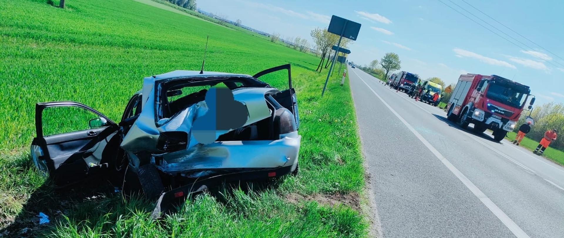 Na zdjęciu widać rozbity srebrny samochód, który stoi na poboczu drogi. Samochód ma rozbity tył oraz otwarte okna. W tle widać stojące na asfaltowej drodze cztery czerwone samochody straży pożarnej oraz żółta karetka pogotowia. Obok pojazdów stoją strażacy oraz ratownicy medyczni. W tle widać drzewa, pachołki oraz znaki drogowe. Jest widno.