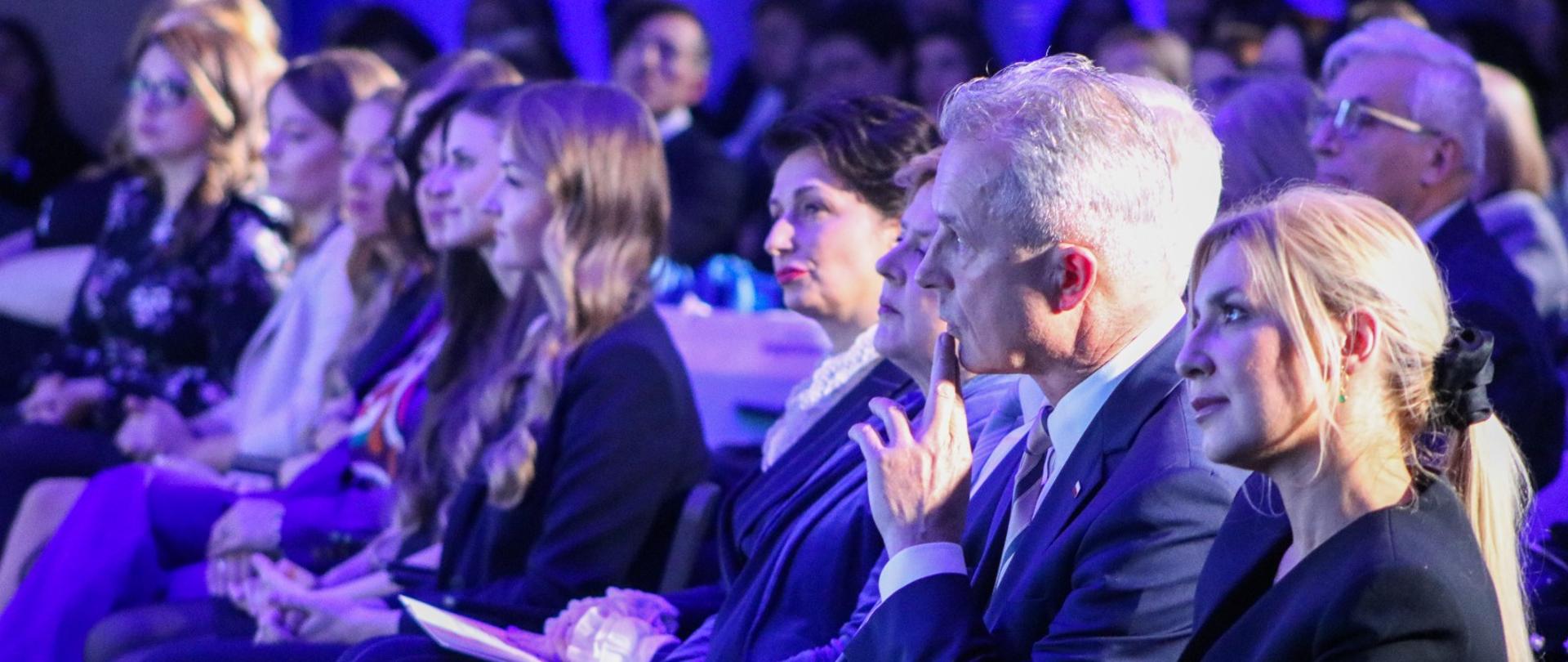 Na oświetlonej na niebiesko sali siedzi wiele osób.