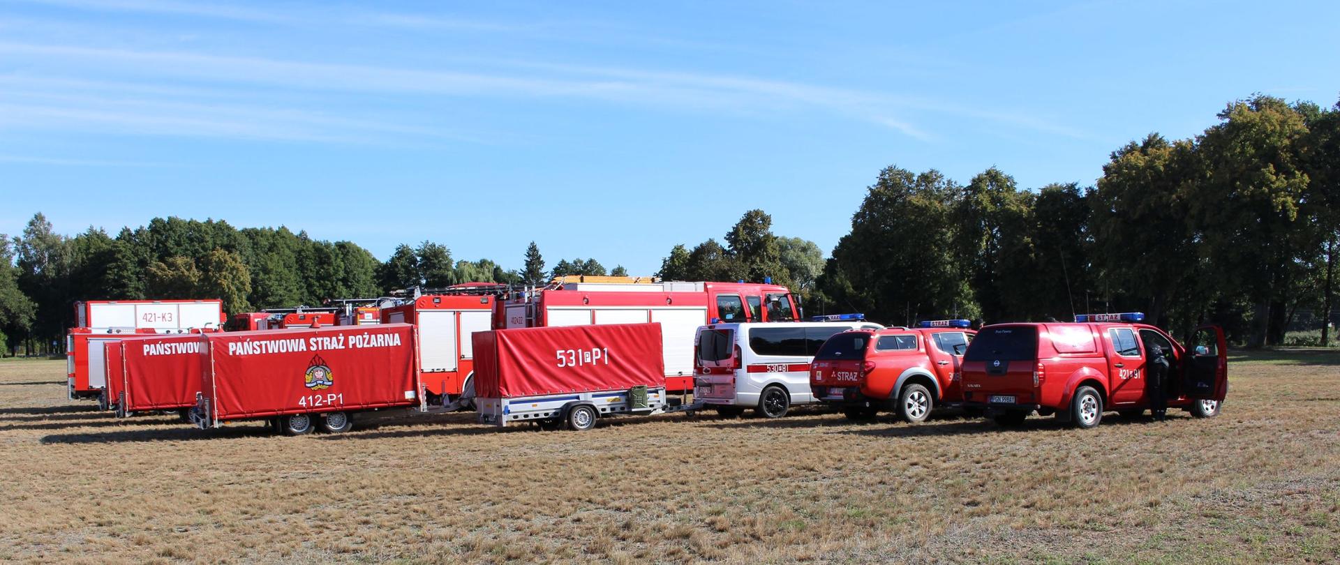 Zdjęcie przestawia strażaków oraz sprzęt strażacki i pojazdy podczas manewrów pod kryptonimem „WODA 2022”.
W tle trawniki i drzewa.
