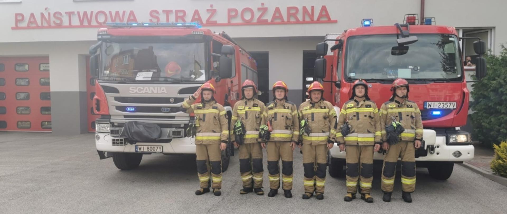 Na zdjęciu widocznych jest sześciu strażaków w piaskowych ubraniach specjalnych i czerwonych hełmach. Funkcjonariusze stoją w szeregu przed samochodami ratowniczo-gaśniczymi, przed bramami sochaczewskiej jednostki.
