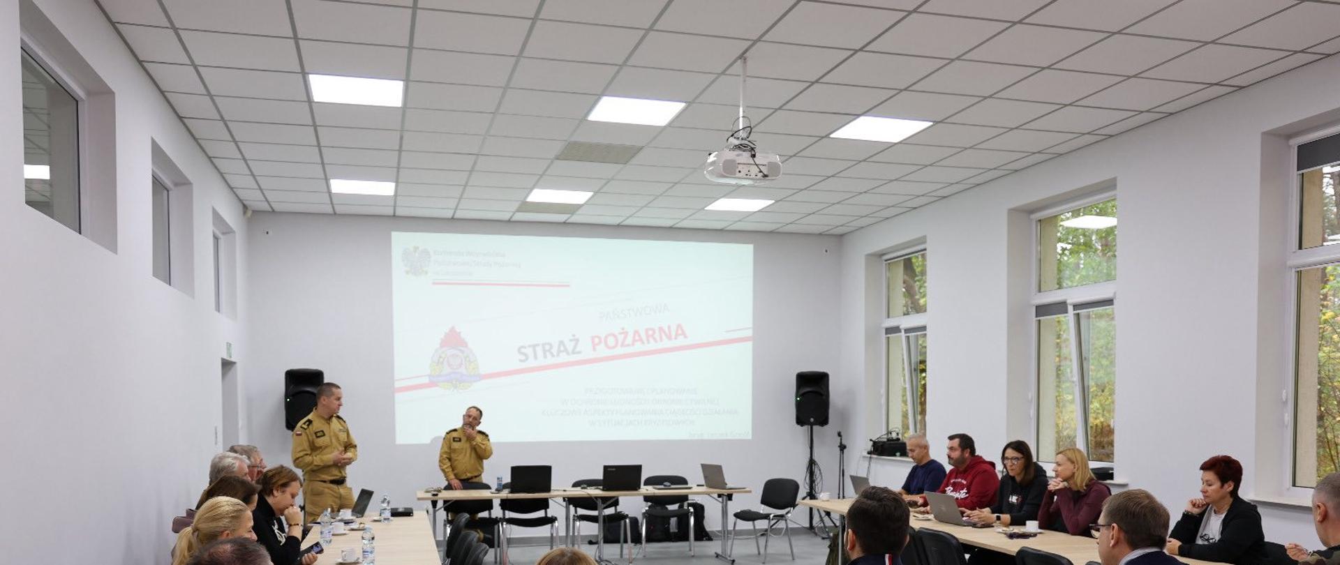 Grupa ludzi siedząca wokół stołu, prowadząca rozmowę. Wszyscy znajdują się w dużej sali, która ma jasno szare ściany i biały sufit.