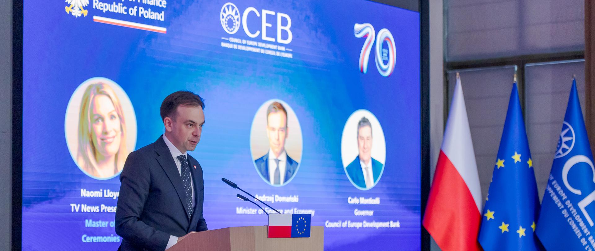 Minister Finansów i Gospodarki Andrzej Domański przy mównicy na tle ekranu z logotypami Ministerstwa Finansów i CEB podczas wydarzenia jubileuszowego. Obok znajdują się flagi Polski, Unii Europejskiej i CEB