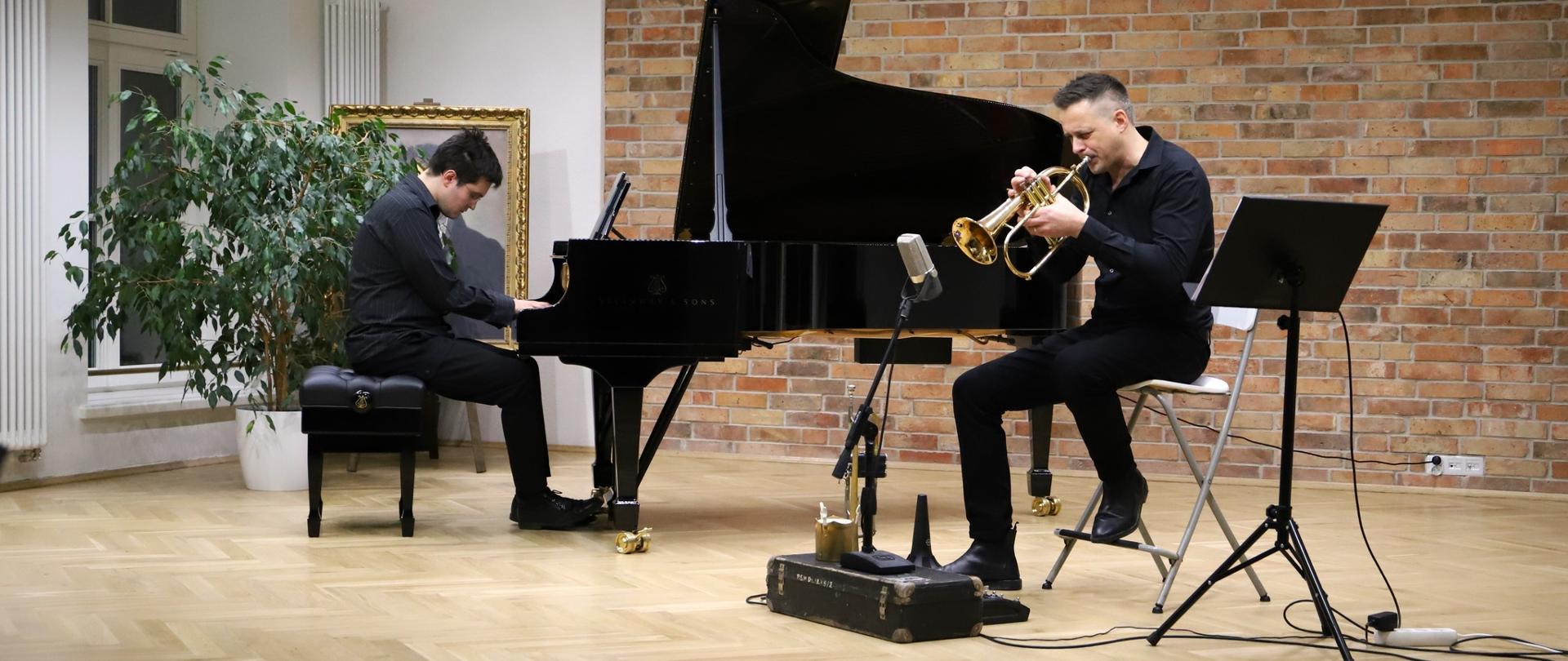 „Dwóch muzyków podczas koncertu w szkolnej auli: po lewej pianista grający na fortepianie, po prawej trębacz siedzący z instrumentem. Obaj występują na jasnej, drewnianej scenie na tle ceglanej ściany, otoczeni roślinami.”