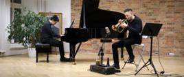 „Dwóch muzyków podczas koncertu w szkolnej auli: po lewej pianista grający na fortepianie, po prawej trębacz siedzący z instrumentem. Obaj występują na jasnej, drewnianej scenie na tle ceglanej ściany, otoczeni roślinami.”