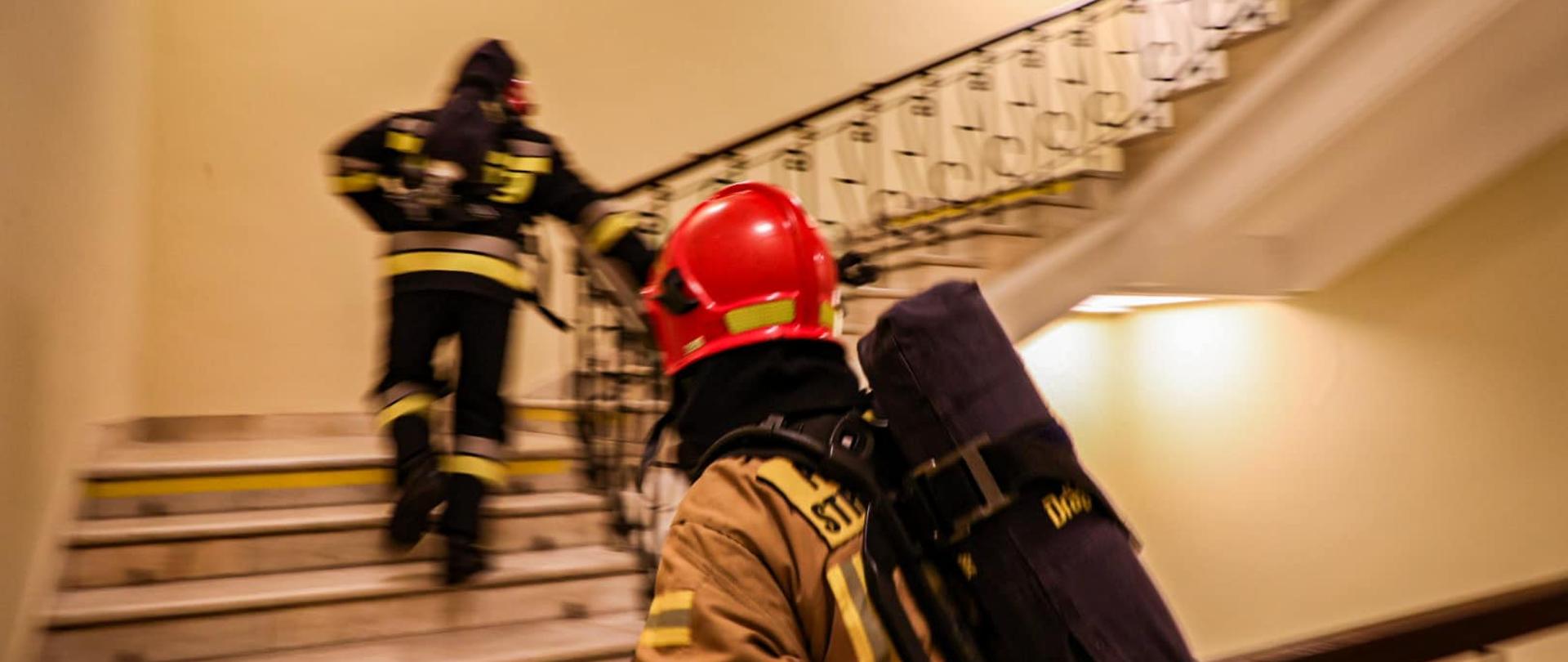 Dwóch strażaków wbiega po schodach po klatce schodowej, jeden za drugim. Obaj trzymają się poręczy. Ubrani są w ubrania specjalne, a na plecach mają założone aparaty powietrzne. 