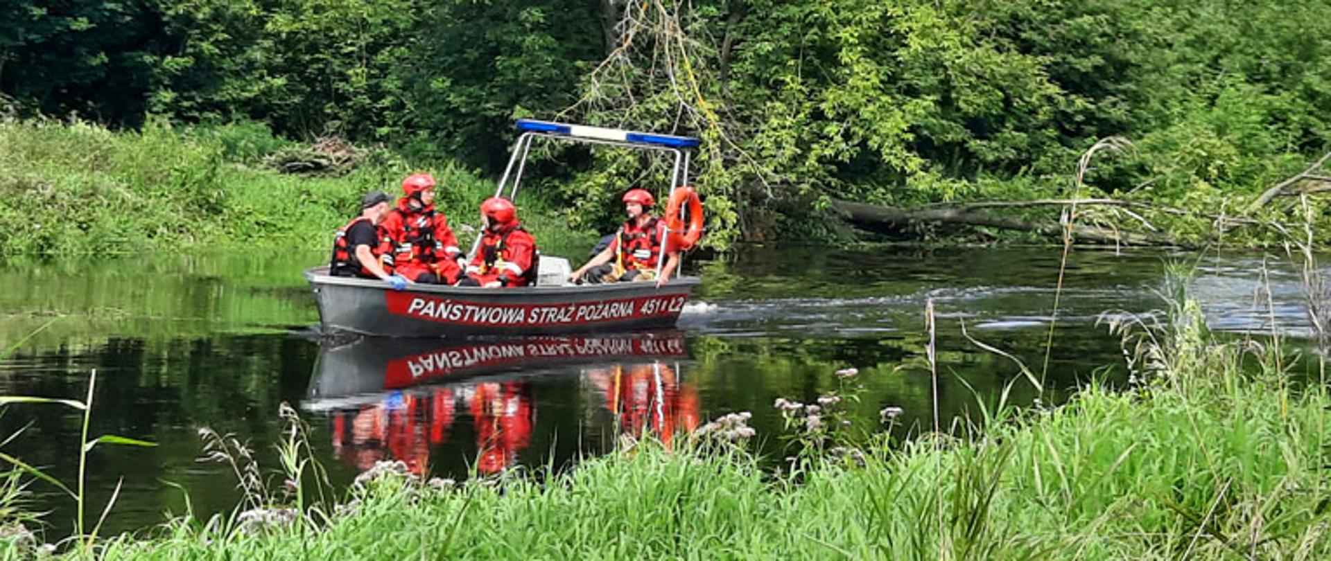 Fotografia przedstawia czterech ratowników siedzących w łodzi płaskodennej aluminiowej, która płynie po rzece, dwóch z nich ma ubrania do pracy w wodzie barwy czerwonej, szelki ratownicze oraz czerwone kaski na głowach, dwóch kamizelki barwy czerwonej z szelkami a jeden z nich ma kask na głowie barwy czerwonej a drugi czapkę z daszkiem. Jeden ratownik siedzi na rufie łodzi i obsługuje silnik motorowy, trzech siedzi na dziobie skierowani twarzami do siebie. Łódź ma zamontowaną belkę ze światłem alarmowym barwy niebieskiej, na jej boku widnieje napis PAŃSTWOWA STRAŻ POŻARNA oraz numer boczny 451EŁ2. Koryto rzeki zarośnięte trawą, dookoła drzewa, świeci słońce.