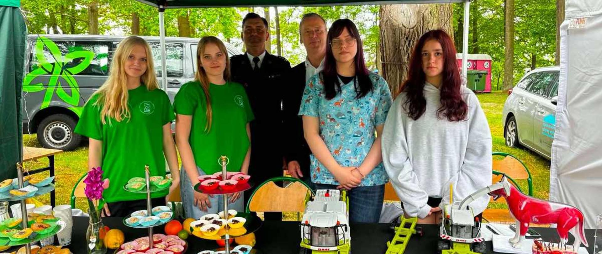 Zdjęcie przedstawia grupę pięciu osób stojących za stołem wystawowym pod zielonym namiotem ogrodowym na świeżym powietrzu.Osoby na zdjęciuDwie młode kobiety po lewej stronie: ubrane w jaskrawozielone koszulki z okrągłym logo na piersi. Obie mają długie, jasne włosy.Mężczyzna w tle (środek-lewo): ubrany w ciemny, oficjalny mundur mundurowy (prawdopodobnie strażacki lub policyjny) z białym kołnierzykiem i krawatem.Mężczyzna w tle (środek-prawo): ubrany w elegancką, ciemną marynarkę i białą koszulę bez krawata.Młoda kobieta po prawej stronie (środek): ma ciemne włosy, okulary i wzorzystą, jasnoniebieską koszulkę medyczną lub weterynaryjną w zwierzątka.Młoda kobieta na skraju po prawej stronie: ma długie, ciemnoczerwone włosy i jest ubrana w luźną, szarą bluzę z kapturem.Stół i eksponatyPrzed ludźmi stoi stół nakryty czarnym obrusem, na którym ułożono:Słodycze: wielopoziomowe patery z babeczkami (muffinkami) w kolorowych papilotkach oraz talerze z ciastkami.Zabawki rolnicze: dwa plastikowe, zielono-białe modele kombajnów lub maszyn rolniczych marki Claas wraz z odczepianym czerwono-żółtym elementem tnącym.Model anatomiczny: mała figurka krowy po prawej stronie, pokazująca z jednej strony umaszczenie, a z drugiej układ mięśniowy lub narządy wewnętrzne.Tło i otoczenieKonstrukcja: nad stołem rozpięty jest duży, zielony dach namiotu imprezowego.Pojazdy: po lewej stronie za ludźmi stoi szary samochód dostawczy z zieloną grafiką w kształcie liścia lub kwiatu. Po prawej stronie widać fragment białego samochodu osobowego.Otoczenie: w głębi tła rosną zielone drzewa, co wskazuje na park, las lub teren piknikowy.