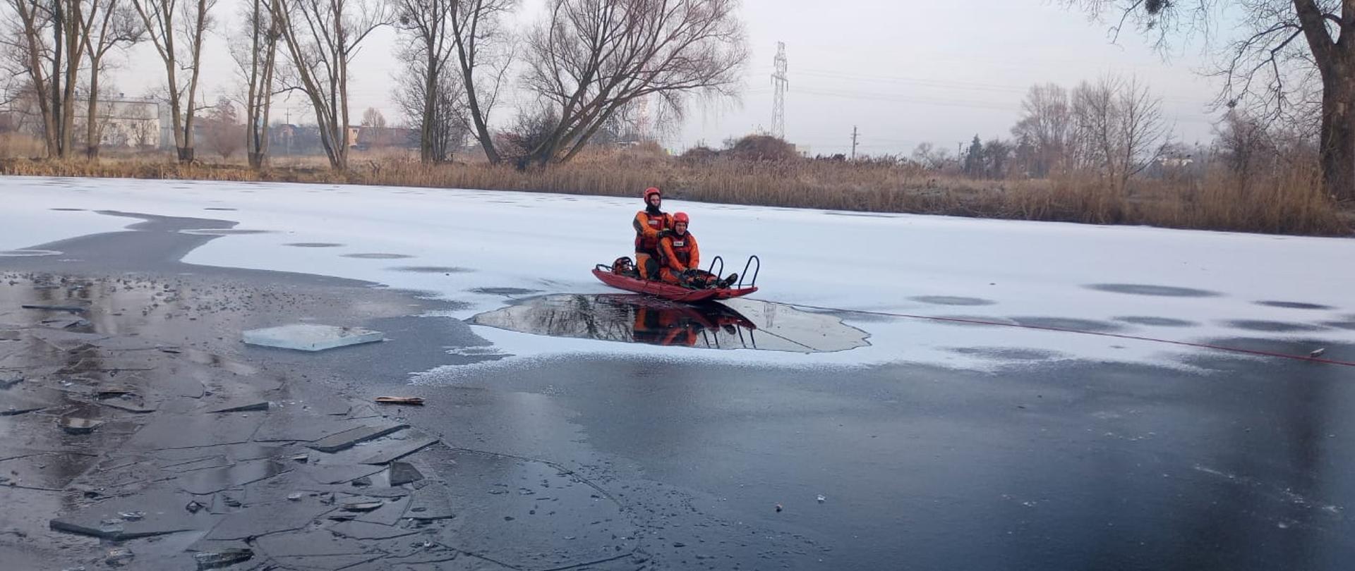 Dwóch ratowników znajduje się na saniach lodowych i przeprowadza ewakuację osoby poszkodowanej z zamarzniętego zbiornika wodnego. Ratownicy zabezpieczeni są suchymi skafandrami, hełmami oraz linką ratowniczą.