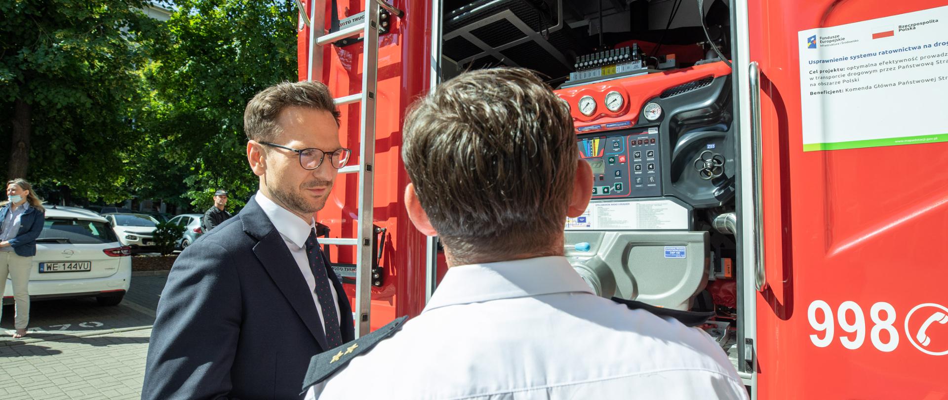 przy wozie Państwowej Straży Pożarnej stoją i rozmawiają dwie osoby - oficer PSP i wiceminister Waldemar Buda
