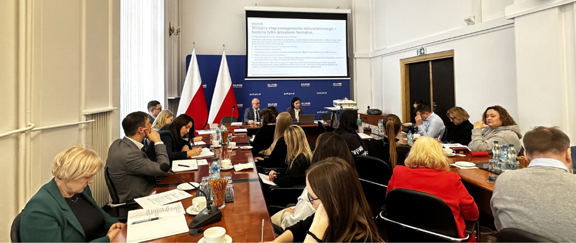 Uczestnicy szkolenia GUNB dla przedstawicieli wojewódzkich inspektoratów nadzoru budowlanego siedzący przy stole konferencyjnym; na ekranie prezentacja dotycząca analizy SWOT prywatyzacji firm państwowych, w tle dwie polskie flagi. 