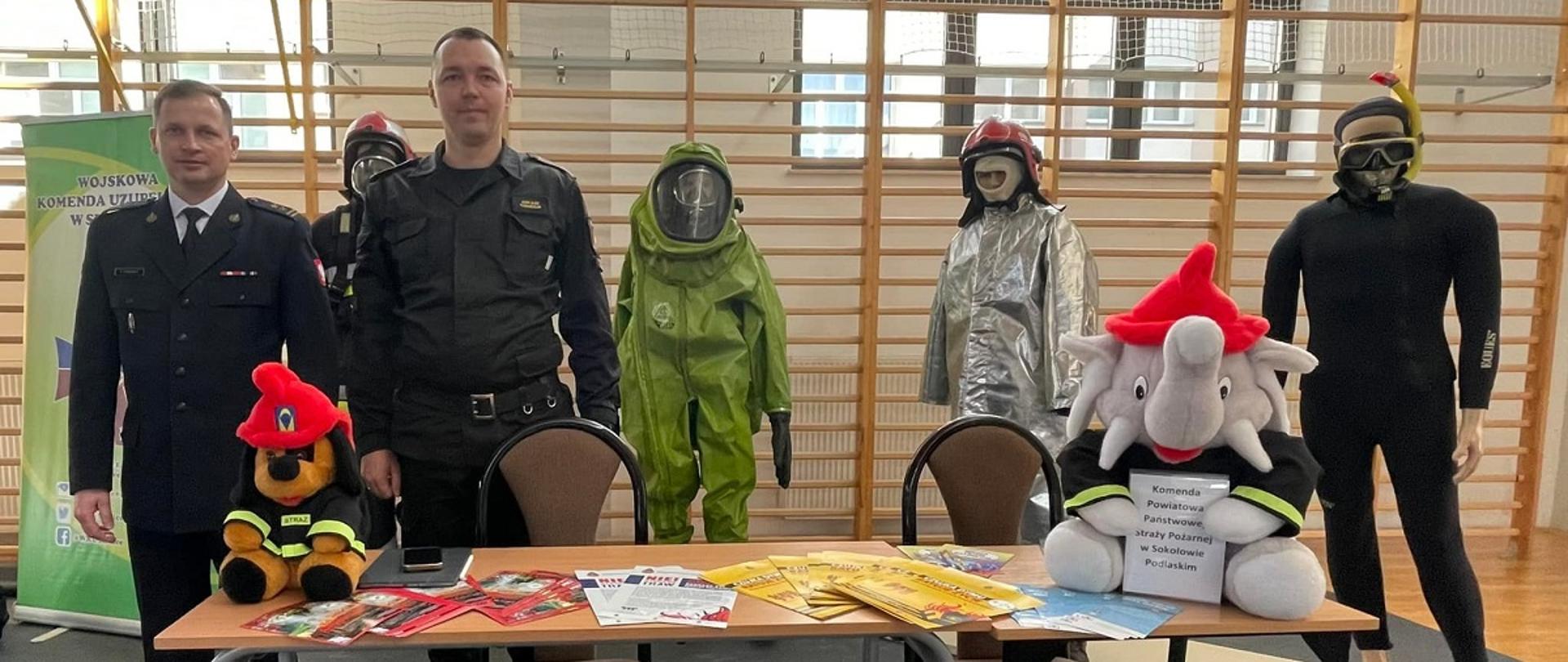XI Sokołowskie Targi Pracy, Edukacji i Kariery „Szarada” - na zdjęciu na sali gimnastycznej 2 strażacy w mundurach, którzy stoją za stoiskiem - stolikiem i 2 krzesłami. Na stole maskotki i artykuły informacyjne. W tle drabinki gimnastyczne i manekiny ubrani w stroje strażackie.