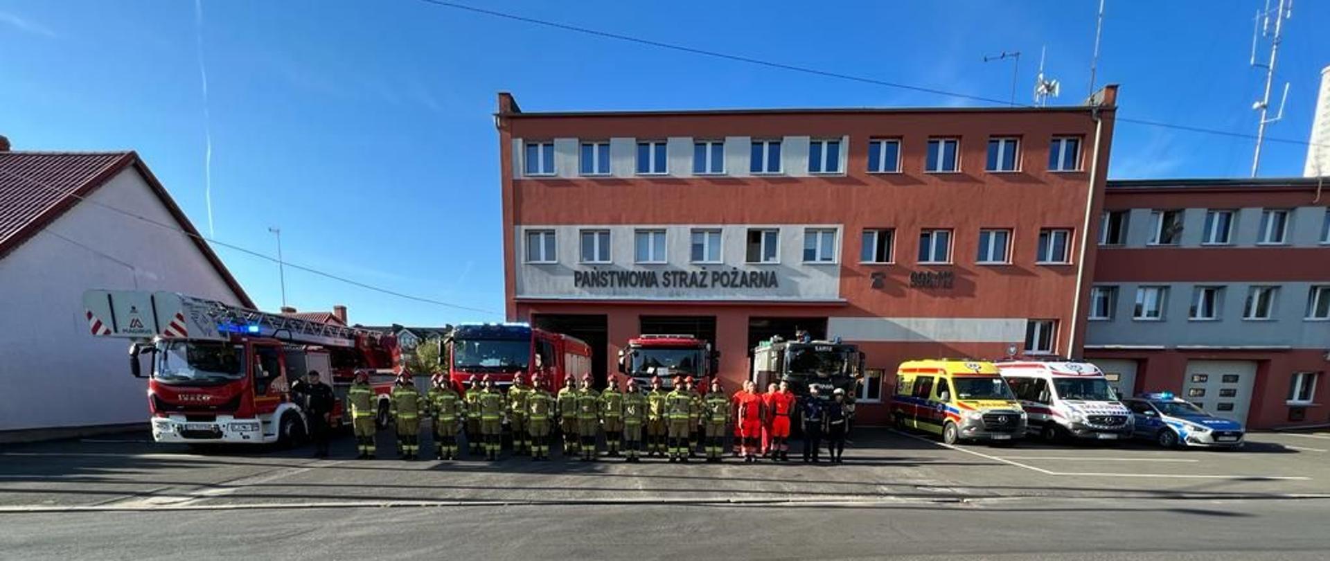 Na tle budynku KP PSP w Nowym Tomyślu stoją wozy strażackie, policyjny radiowóz i karetki pogotowia ratunkowego, na tle samochodów stoją strażacy, ratownicy medyczni i policjanci. Strażak pierwszy od lewej oddaje honor.