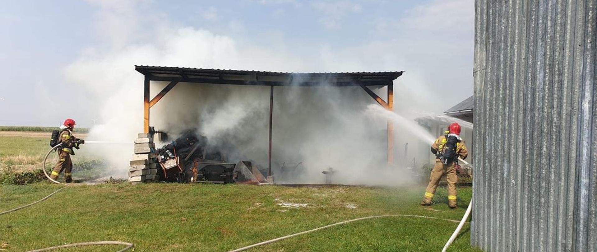 Dwóch strażaków ubranych w aparaty ochrony układu oddechowego podaje prądy wody na palącą się wiatę