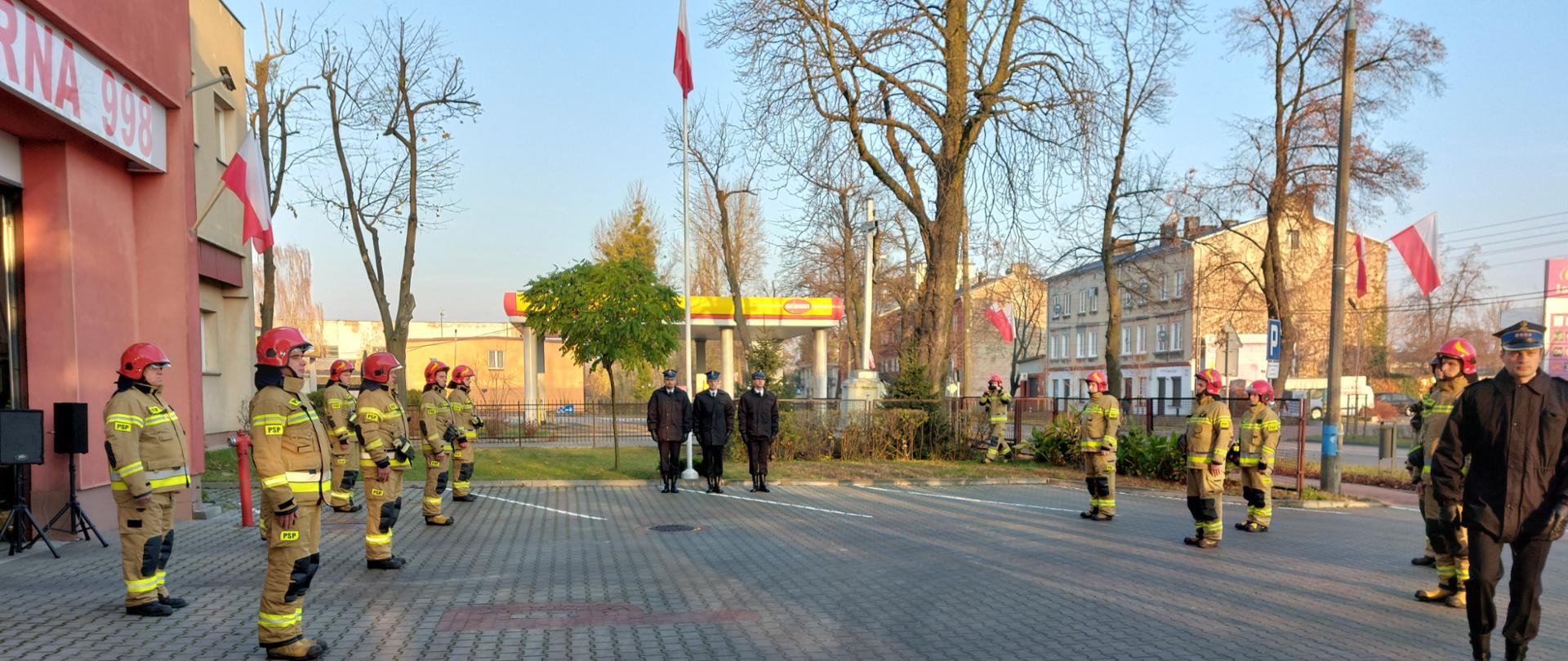 Na placu zewnętrznym komendy stoi przed masztem z flagą poczet flagowy. Po obu stronach stoją strażacy w umundurowaniu bojowym w dwóch szeregach.
