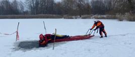 Strażacy podczas ćwiczeń ratownictwa wodno-lodowego na zamarzniętym akwenie.
Dwóch strażaków w pomarańczowych kombinezonach ratowniczych na zamarzniętym jeziorze podczas ćwiczeń; jeden leży na saniach lodowych w pobliżu przerębla, a drugi go holuje.