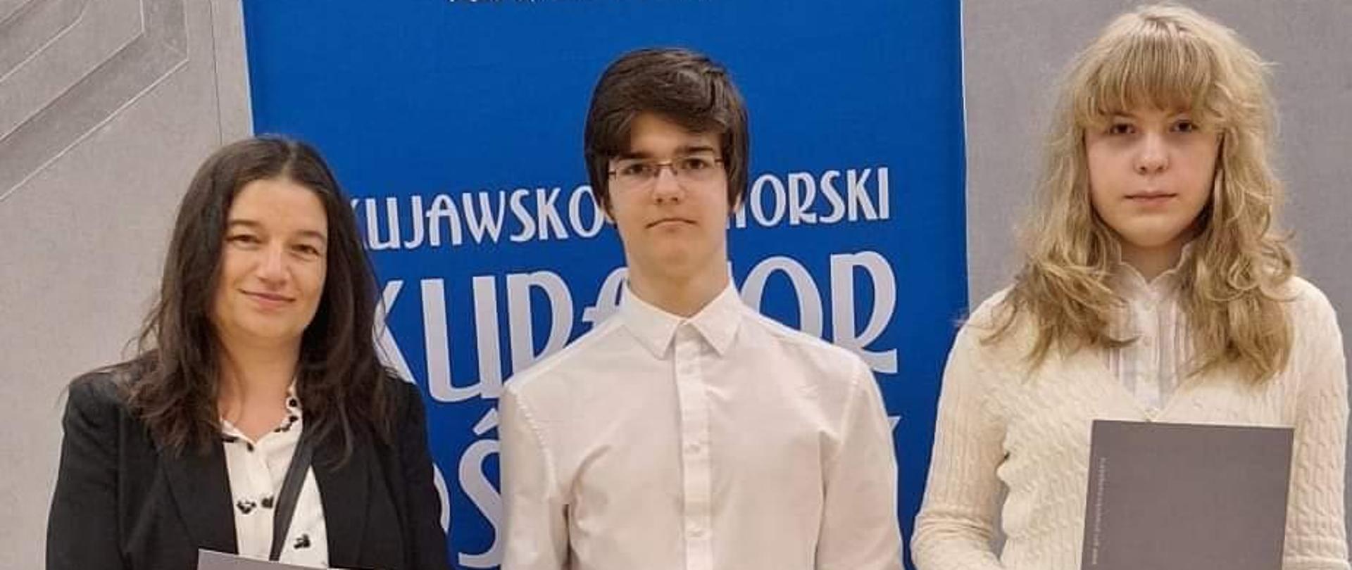  po lewej chłopak w okularach i czarnych włosach po prawej dziewczyna w blond włosach białej koszuli i szarych spodniach w tle niebieski ekran z napisem kuratorium oświaty . Oboje trzymają w rękach szare teczki z dyplomem towarzyszy im na zdjęciu nauczycielka również z szarą teczką