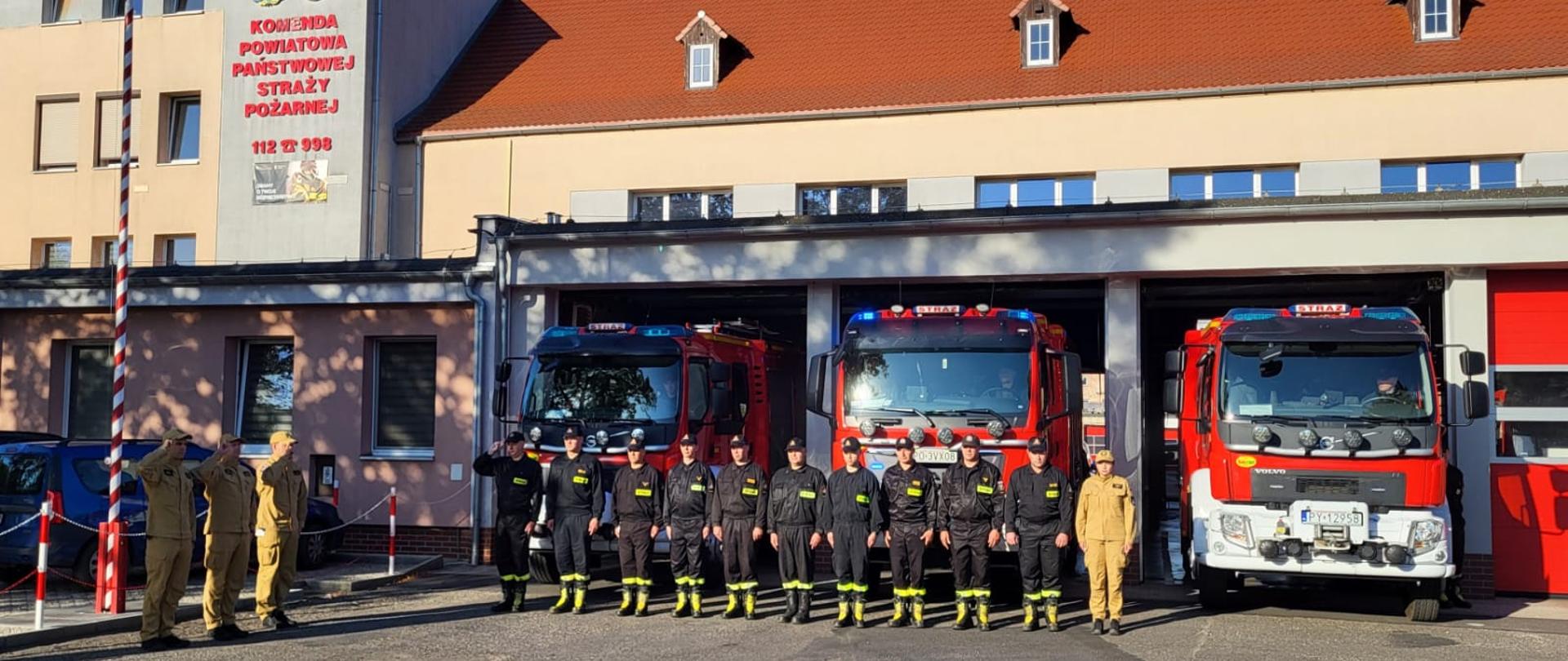Na zdjęciu widać strażaków przed budynkiem Komendy, samochody strażackie wyjechały z garażu, mają włączone sygnały świetlne i dźwiękowe, widać maszt z flagą Polski i budynek Komendy Powiatowej PSP w Kościanie.