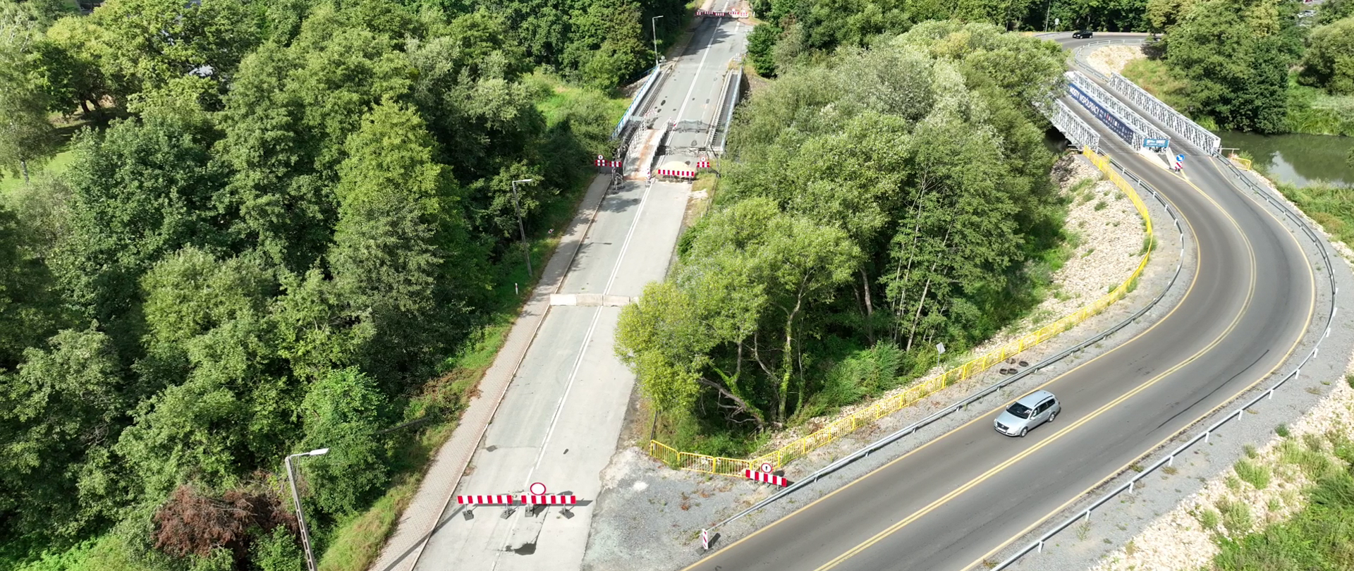 Fragment DK45 w Krapkowicach po powodzi. W okolicy obszar z rzeką przepływającą przez środek kadru, otoczony gęstą zielenią (drzewa i krzewy). Nad rzeką Osobłoga znajdują się dwa mosty - zamknięty - po powodzi oraz most tymczasowy, którym odbywa się ruch.
