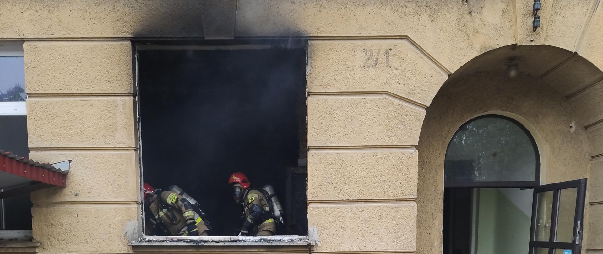 Zdjęcie przedstawia budynek opuszczonego szpitala. W oknie widać dwóch strażaków podczas akcji gaśniczej. Z okna wydobywa się dym, a ściana powyżej jest okopcona. Drzwi po prawej stronie są otwarte i widać wąż gaśniczy rozłożony na schodach. Pod oknem leżą nadpalone elementy wyrzucone z gabinetu. 