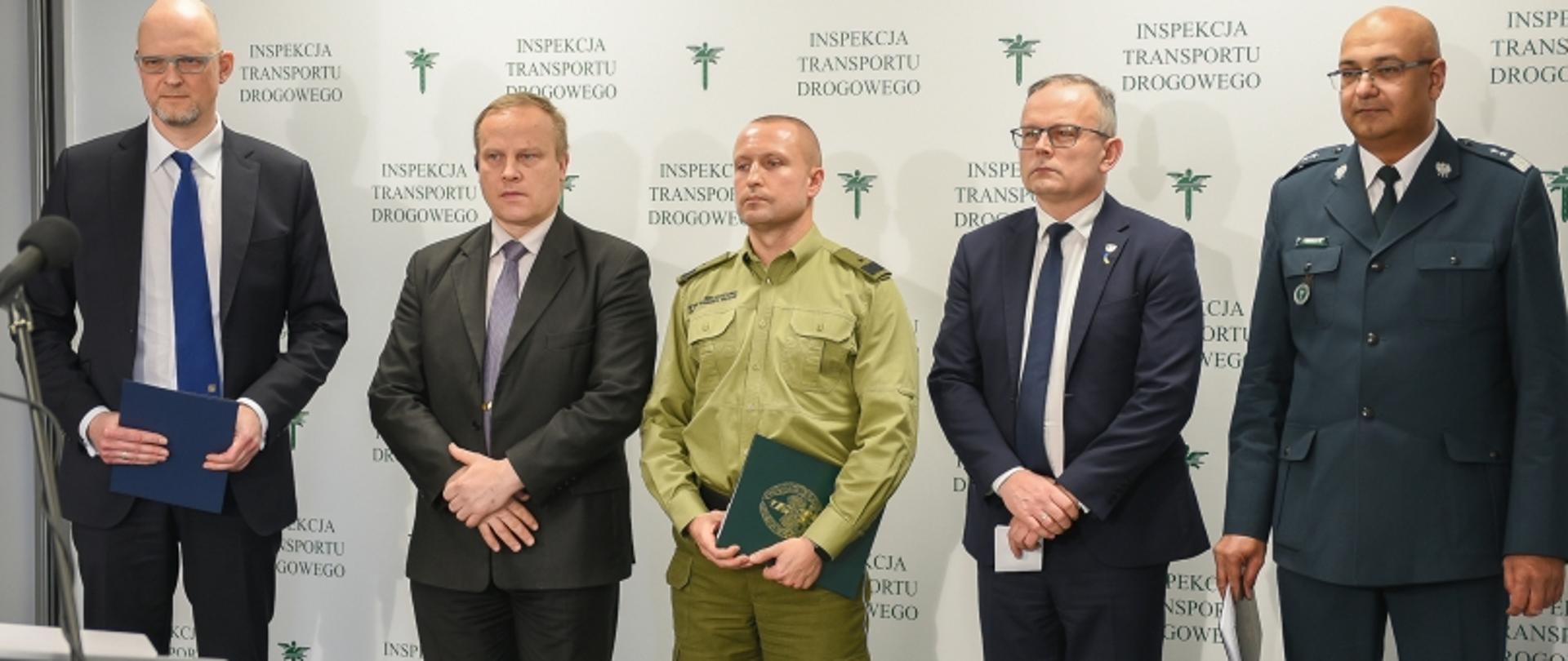 Pięć osób na tle ścianki „INSPEKCJA TRANSPORTU DROGOWEGO”, w tym osoby w mundurach; po lewej mikrofon na statywie.