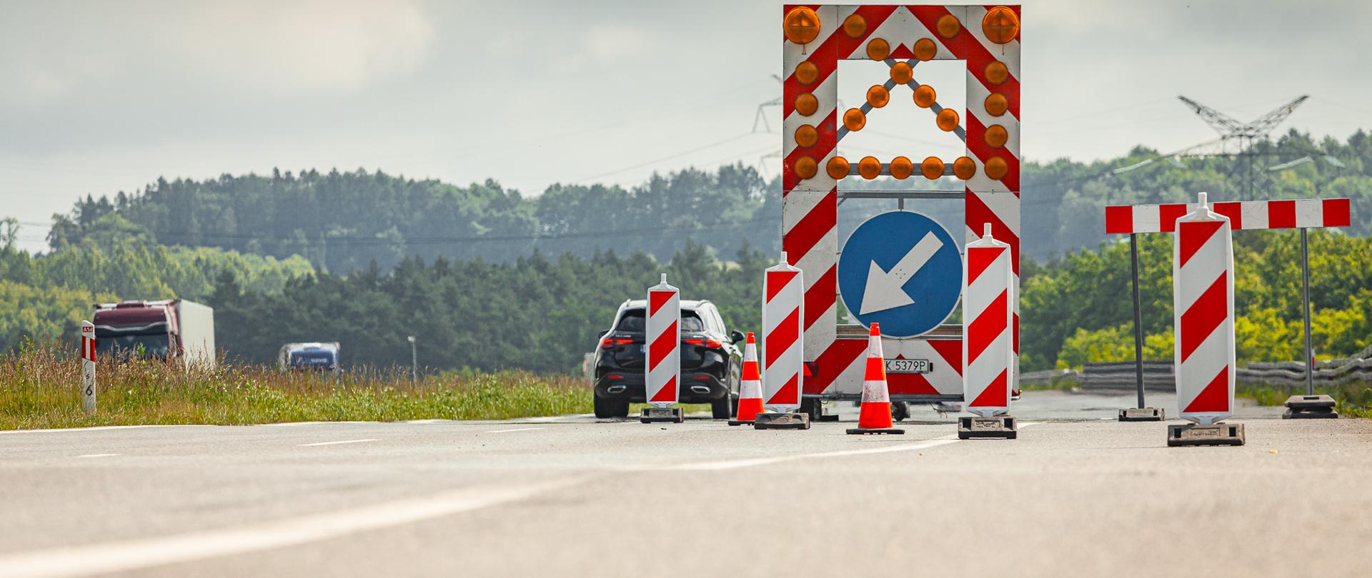 Zdjęcie przedstawia tymczasową organizację ruchu na drodze podczas robót drogowych. Na środku jezdni ustawiono dużą tablicę prowadzącą z czerwono-białymi pasami, migającymi światłami ostrzegawczymi i niebieskim znakiem nakazującym ominięcie przeszkody z lewej strony. Przed tablicą oraz obok niej znajdują się pomarańczowe pachołki i czerwono-białe słupki kierujące ruch.
Po lewej stronie widoczny jest samochód osobowy oraz ciężarówka poruszające się wyznaczonym pasem. W tle rozciąga się pas lasu, linie energetyczne i zachmurzone niebo. Fotografia wykonana została z niskiej perspektywy, podkreślającej szerokość jezdni i zabezpieczenia w miejscu prowadzonych prac drogowych.