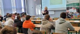Pracownica KRUS stojąc przed uczniami przeprowadza wykład. Uczniowie siedzą w szkolnych ławkach i z uwagą słuchają przekazywanych im informacji.