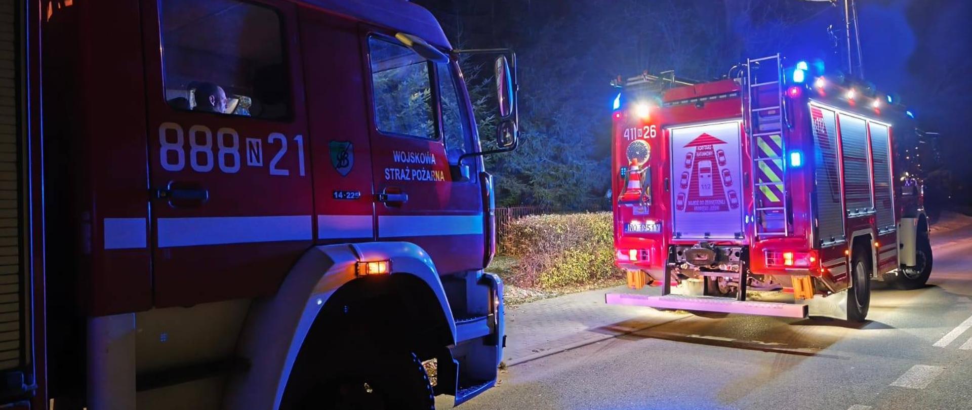 Zdjęcie zrobione nocą. Na drodze stoją dwa samochody strażackie na niebieskich światłach ostrzegawczych.