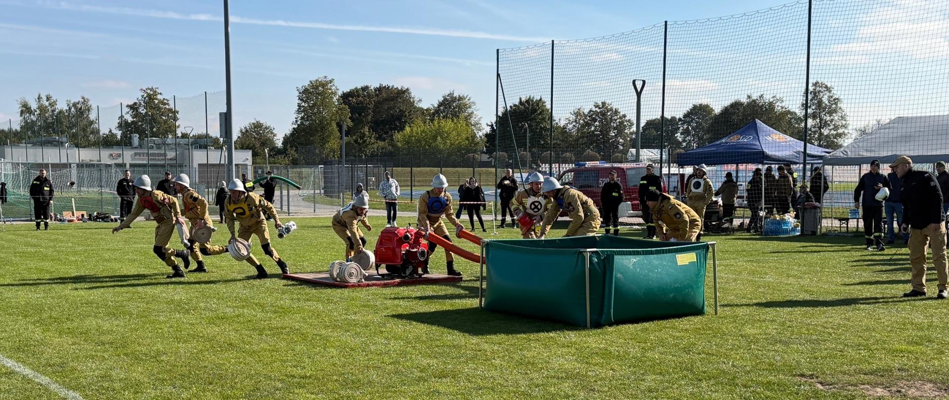 Drużyna OSP w trakcie wykonywania ćwiczenia bojowego. Strażacy rozwijają węże i uruchamiają motopompę przy zbiorniku wodnym. W tle obecni sędziowie i kibice.