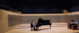 Nastolatek gra na fortepianie w sali koncertowej. Po prawej stronie estrady stoją organy.