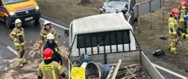dwa samochody rozbite srebrny stoi na poboczy drogi biały typu bus na drodze po lewej stronie wysypany na drodze sorbent oraz stoi czterech strażaków na pace samochodu bus materiały budowlane u góry karetka pogotowia