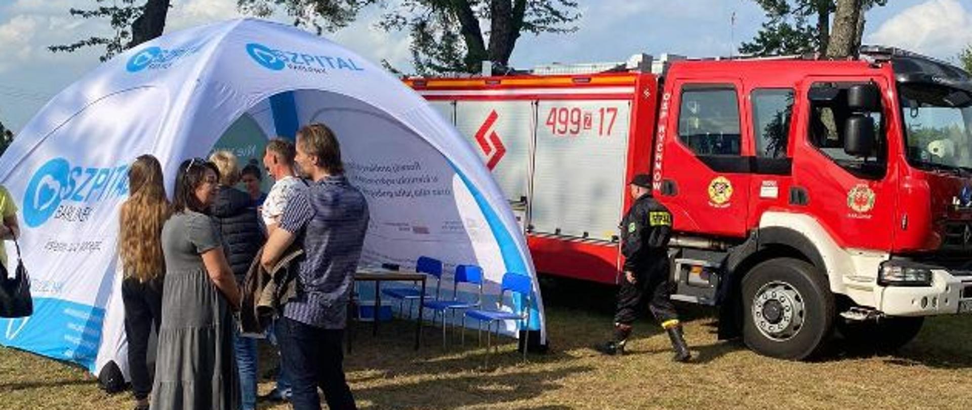 Na zdjęciu osoby oczekujące na zaszczepienie się w punkcie szczepień - namiocie. Obok stoi samochód strażacki.