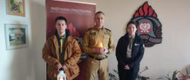 Na zdjęciu znajdują się strażak w mundurze służbowym oraz dwoje harcerzy w mundurach, trzymają zapalony lampion