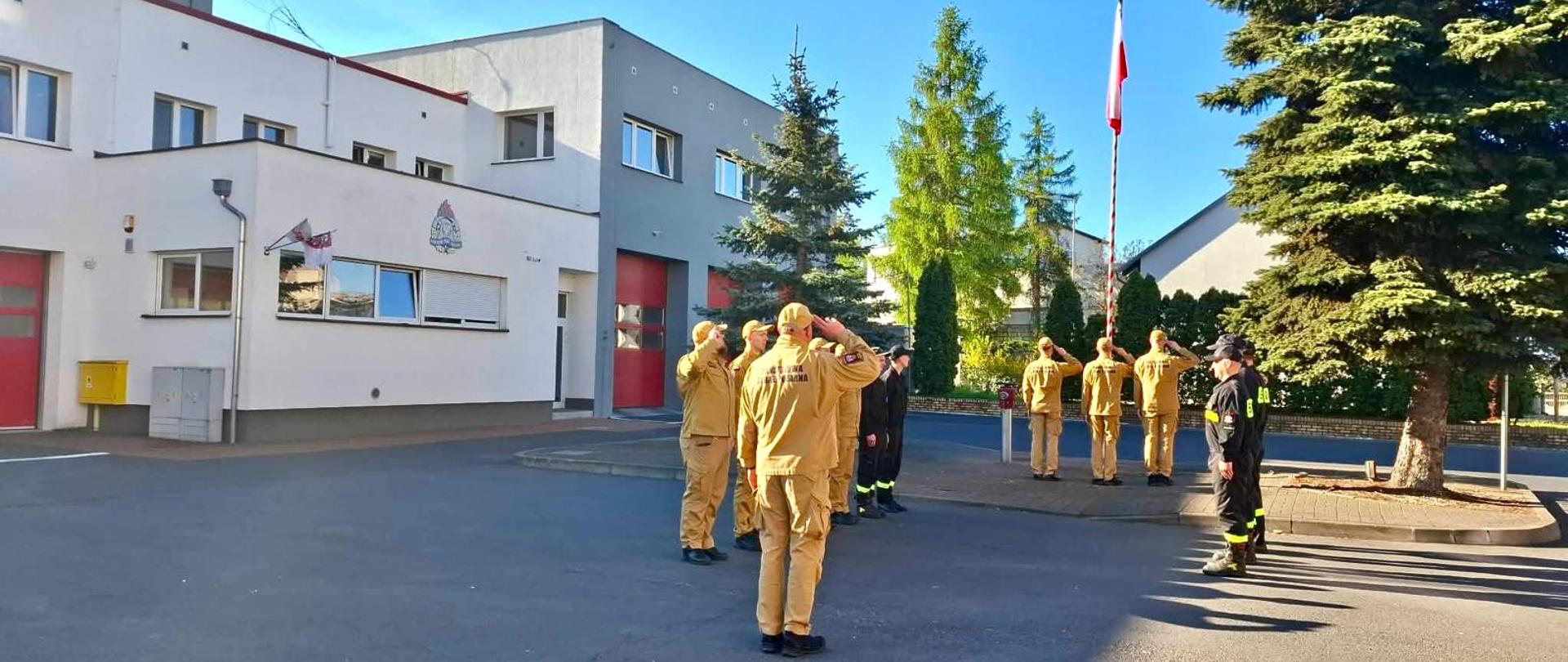 Grupa umundurowanych osób stoi przy wysokim maszcie na chodniku wzdłuż ulicy. Jedna osoba trzyma biało-czerwoną flagę, przygotowując ją do wciągnięcia. W tle widać niskie budynki mieszkalne, drzewa oraz zaparkowany samochód. Jest słoneczny poranek, niebo jest bezchmurne.