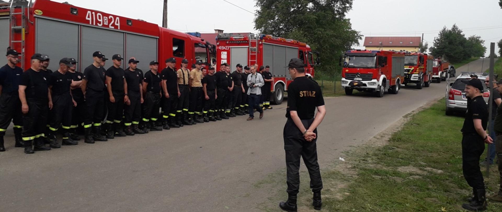Kolorowe zdjęcie wykonane w przestrzeni otwartej w dzień. Przedstawia wielu umundurowanych strażaków na zbiórce w dwuszeregu, w koszulkach z krótkim rękawem, z czapkami na głowach, na tle ustawionych czerwonych pojazdów ratowniczo-gaśniczych. Strażacy i pojazdy stoją na drodze a w ich kierunku przemawia stojący naprzeciw umundurowany strażak Państwowej Straży Pożarnej. W tle widoczne drzewa i zabudowania a obok po prawej stronie przemawiającego stoją inne osoby umundurowane i cywilne biorące udział w ćwiczeniach.