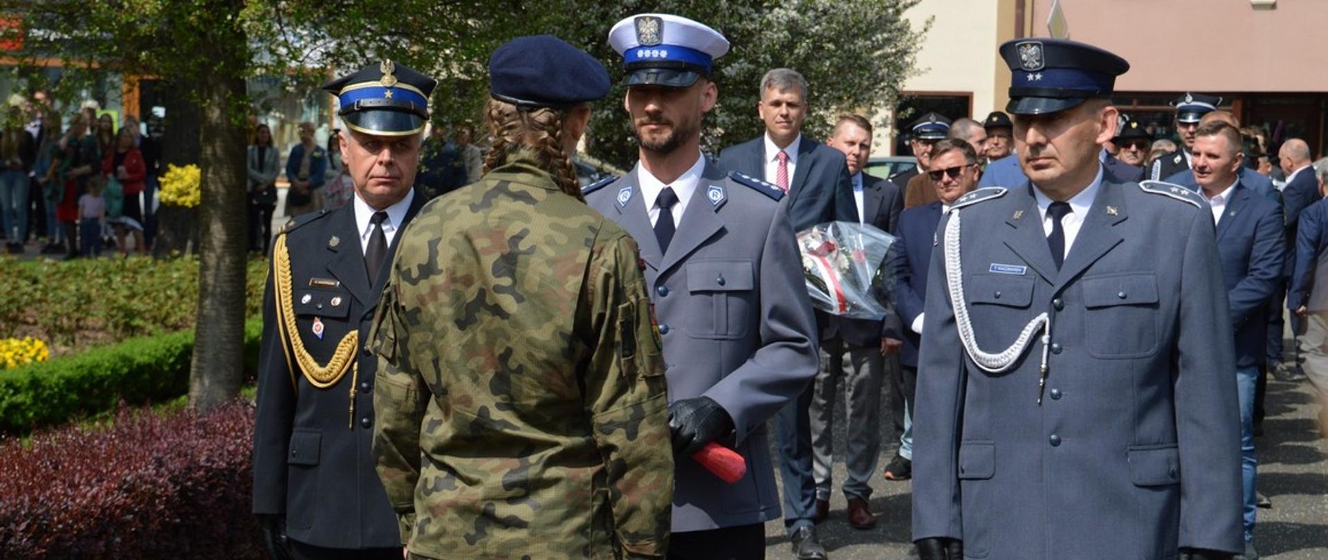 Narodowe Święto Trzeciego Maja. Plac Wolności w Rawiczu. Delegacja służb mundurowych składa wiązankę kwiatów. W skład delegacji wchodzi strażak PSP, stojący z lewej strony, policjant w białej czapce - w środku oraz funkcjonariusz Służby Więziennej - z prawej strony. Przed nimi uczennica klasy mundurowej, która odbiera wiązankę kwiatów. W tle inne delegacje i budynki.