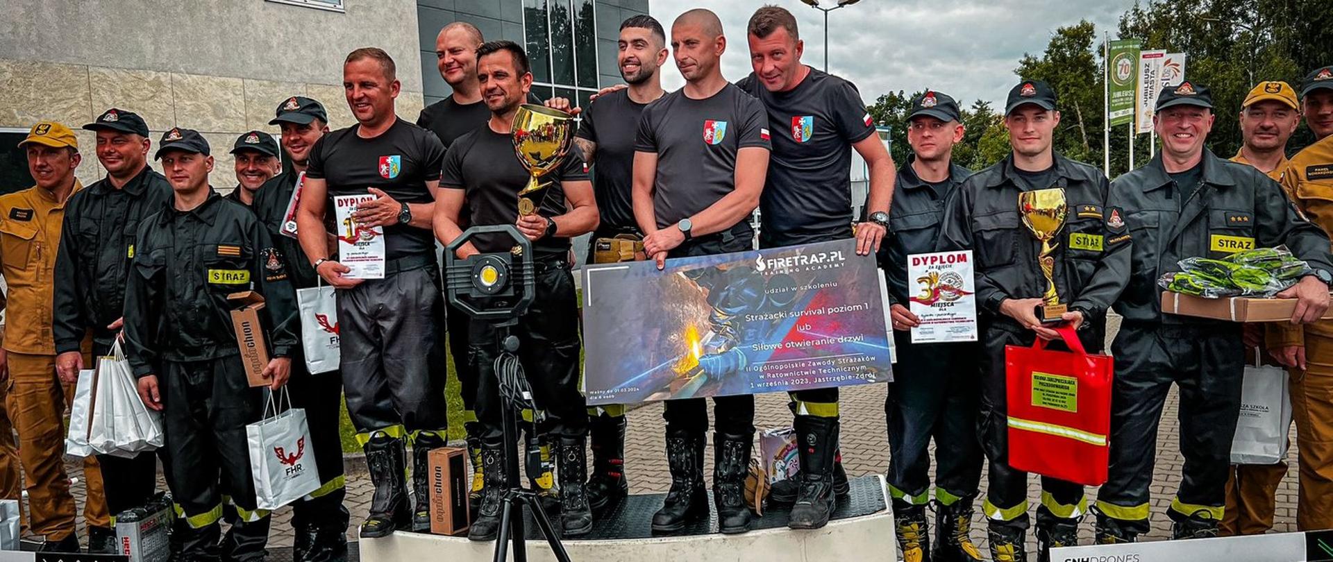 Zdjęcie zrobione na zewnątrz, podczas wręczania nagród strażakom biorącym udział z w zawodach. Na podium stoją zwycięskie drużyny. 
