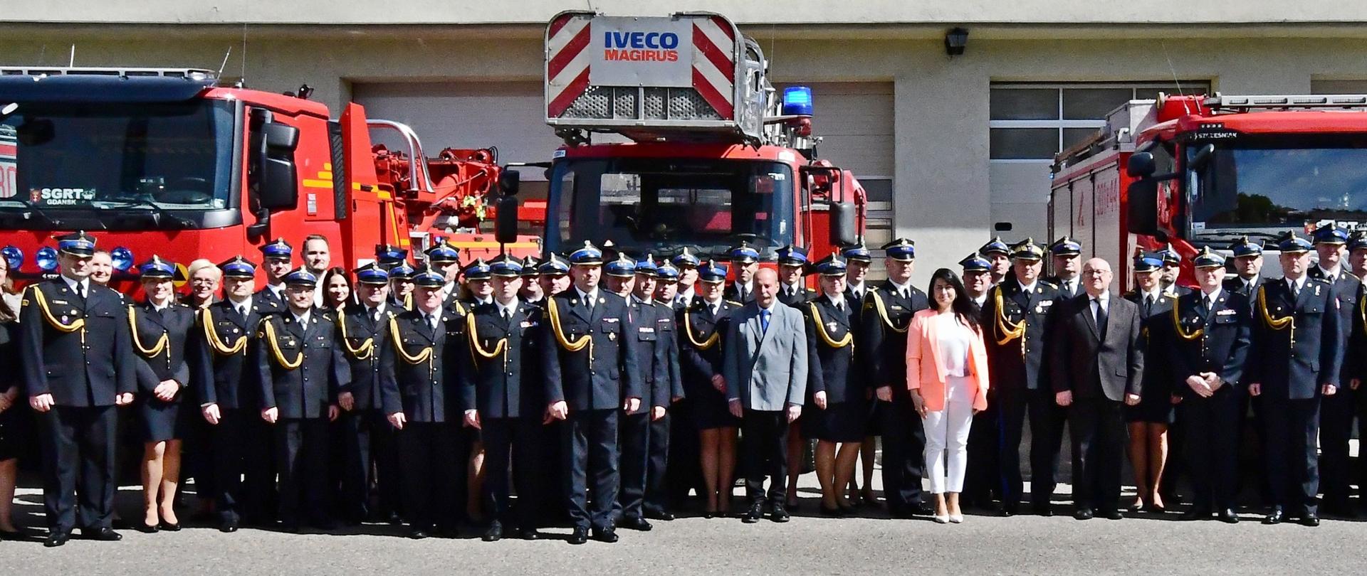 Strażacy oraz pracownicy cywilni stoją obok siebie na tle samochodów gaśniczych.