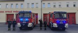 Przed budynkiem Jednostki Ratowniczo-Gaśniczej stoją dwa wozy strażackie z włączonymi niebieskimi światłami. Na tle remizy ustawionych jest sześciu strażaków w mundurach galowych i bojowych, z których trzech oddaje honory.