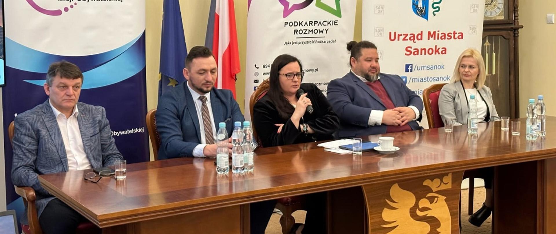 Wiceminister funduszy i polityki regionalnej Małgorzata Jarosińska-Jedynak na spotkaniu z samorządowcami i przedsiębiorcami w Sanoku, wiceminister siedzi za stołem i przemawia przez mikrofon, na nią flagi Polski, UE