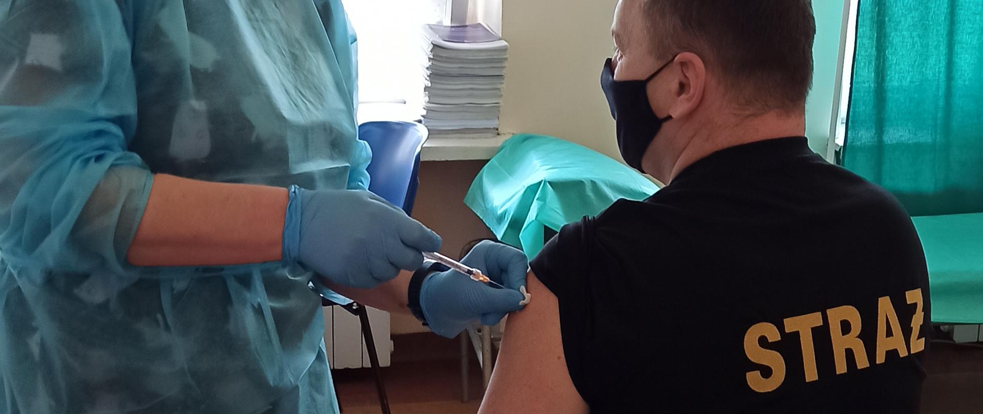 Zdjęcie przedstawia mężczyznę siedzącego na krześle z odsłoniętym ramieniem, który jest szczepiony, przez pielęgniarkę. 