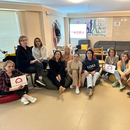 11 kobiet siedzi w półkolu na podłodze i na krzesłach i pozuje do zdjęcia; dwie trzymają plakaty z logo Polskiej pomocy