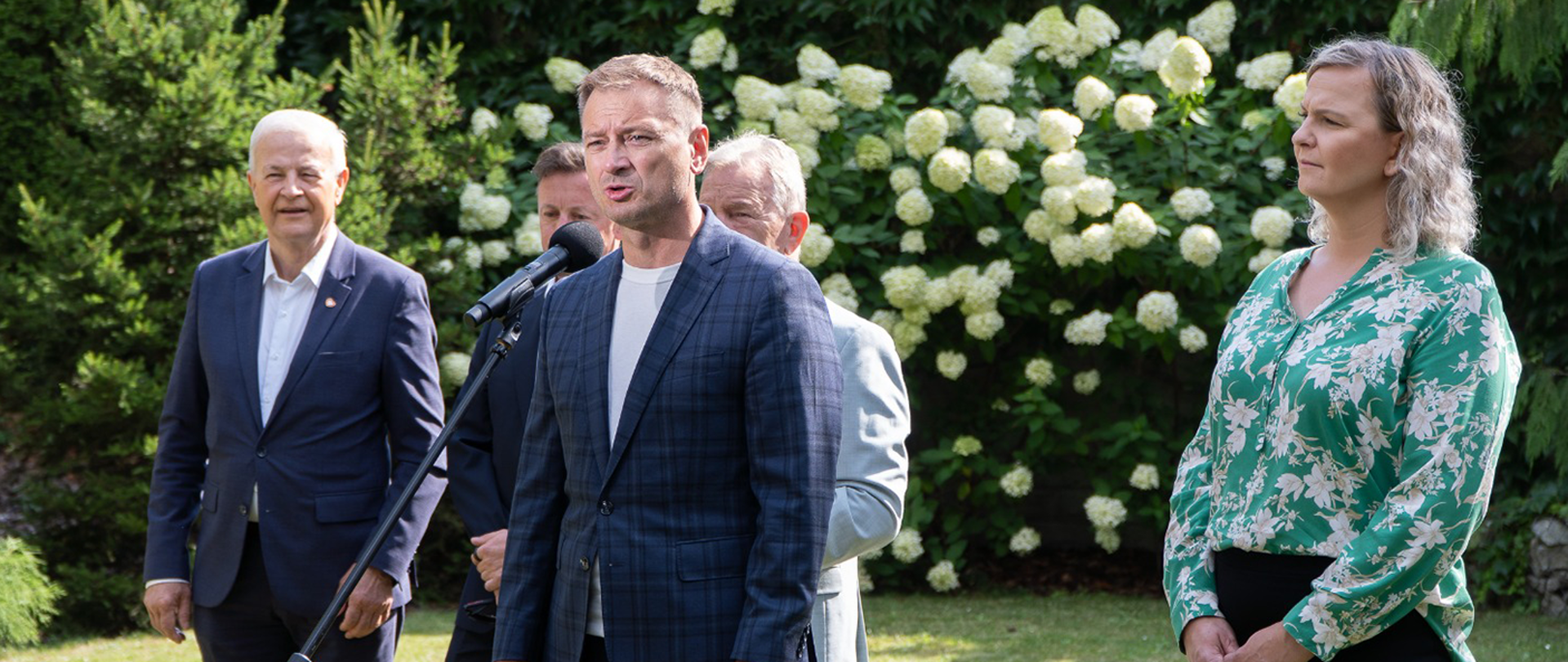 Konferencja prasowa Ministra Sportu i Turystyki Sławomira Nitrasa w ogrodach Sejmu. Minister Nitras w granatowej marynarce w kratę mówi do mikrofonu. Z prawej strony zdjęcia stoi Otylia Jędrzejczak. Z lewej strony stroją Apoloniusz Tajner i częściowo schowani za ministrem Adam Małysz i Andrzej Supron. W tle zielona roślinność i białe kwiaty.