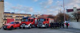 Trzy samochody pożarnicze stoją obok siebie strażacy ustawieni w rzędzie stoją obok siebie poczet sztandarowy wciąga flagę Polski na maszt.